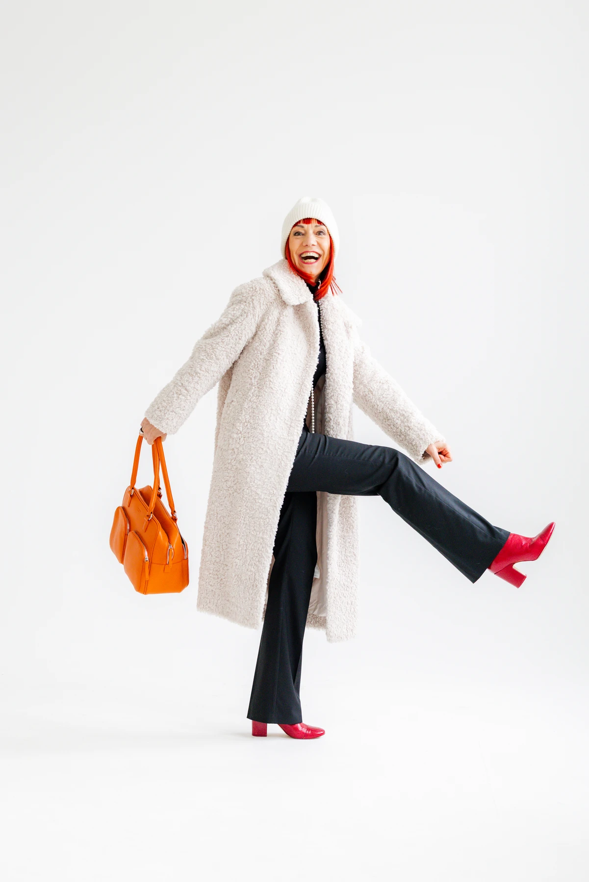 Rothaarige Unternehmerin im cremefarbenen Mantel mit orangefarbener Tasche und roten Stiefeln