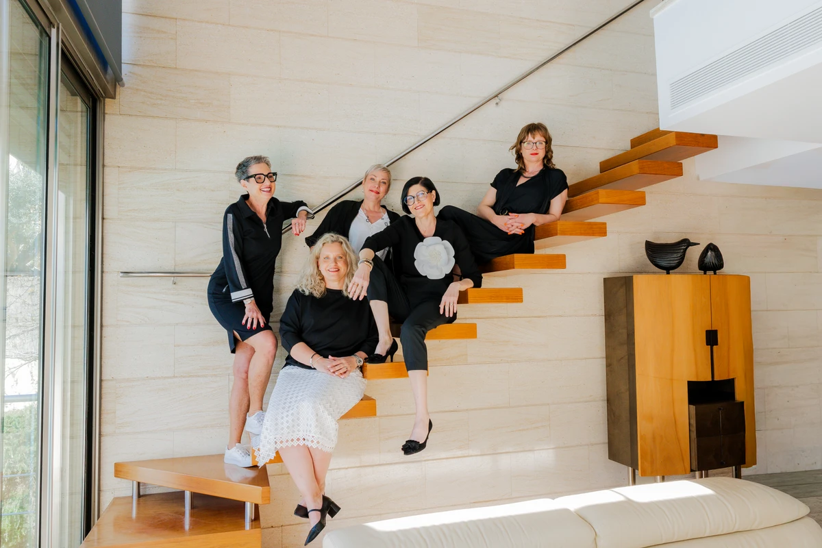 Fünf Frauen in schwarzen Outfits stehen auf einer Holztreppe vor einer cremefarbenen Wand